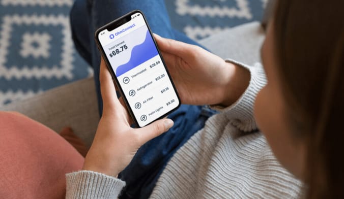 A woman sits on a couch looking at a smartphone with a message on energy savings from company OhmConnect