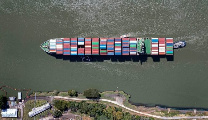 An aerial image of a cargo ship piled with colorful containers sailing in a waterway
