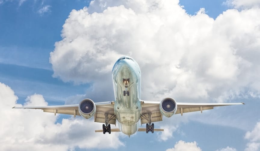 A silver plane flies in the sky against a backdrop of a light blue sky and fluffy white clouds