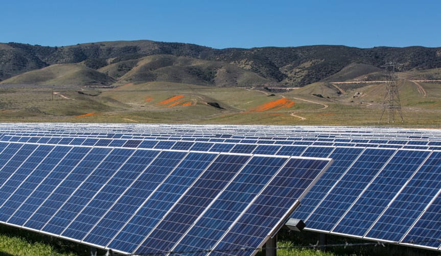 Many rows of solar panels in the foreground, low rolling hills bedecked with smatterings of orange poppy fields in the back