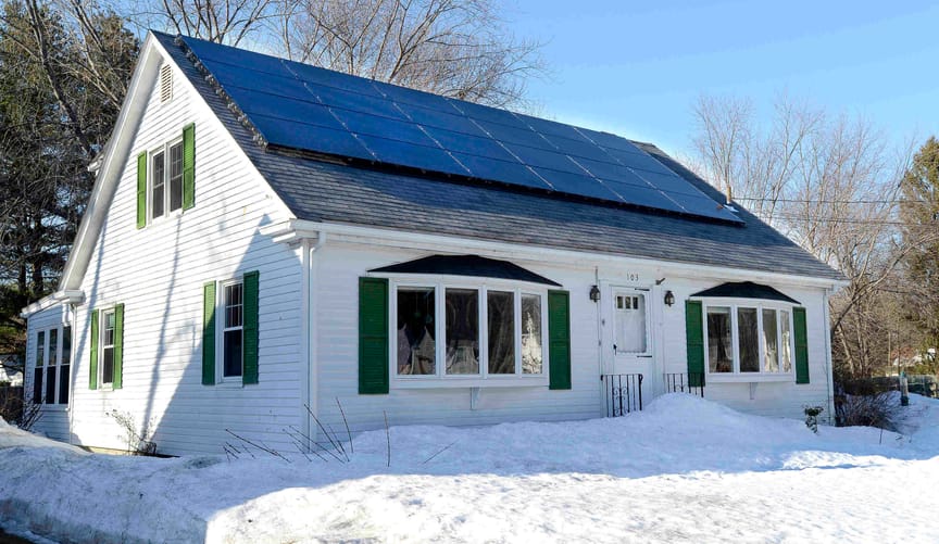 A white bungalow with green shutters and solar panels on the roof. The front yard is covered in snow.
