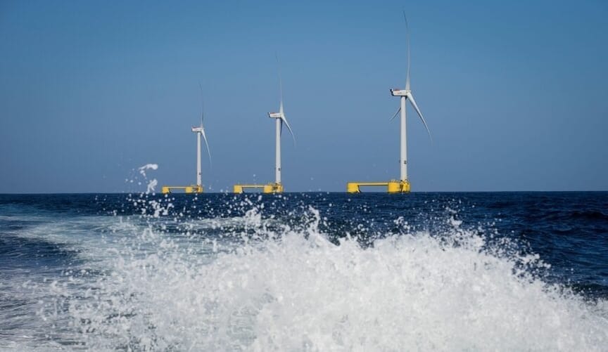 A row of white floating offshore wind turbines in the ocean; in the foreground is a frothy white wave
