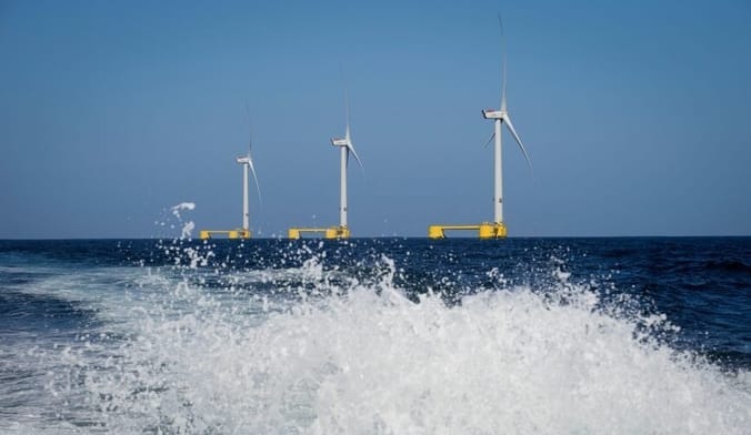 A row of white floating offshore wind turbines in the ocean; in the foreground is a frothy white wave