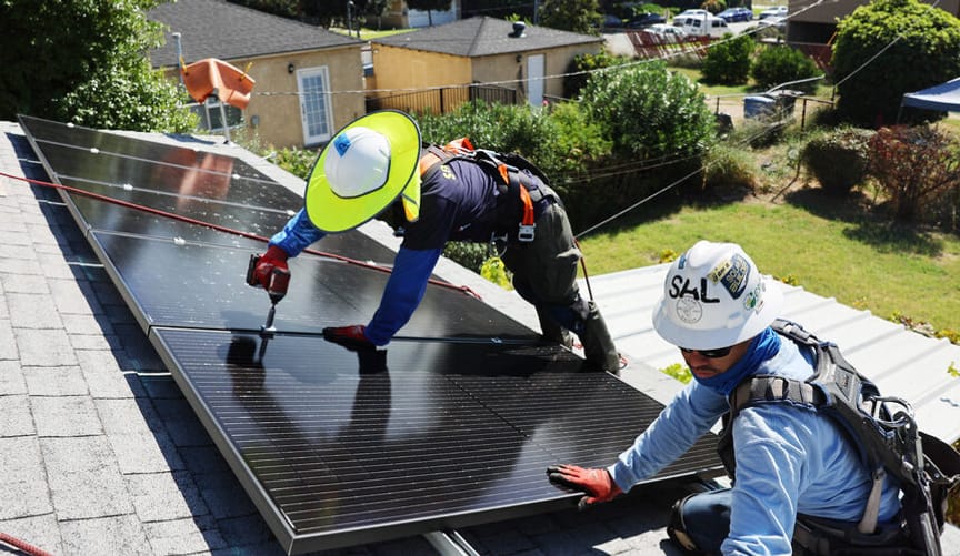 Two workers wearing safety gear install solar panels on the roof of a home
