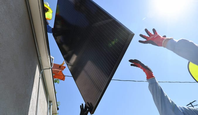 Several workers in safety gear hoist a solar panel onto the roof of a home