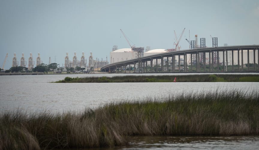In the distance across a river and behind an arching bridge sits an enormous industrial facility
