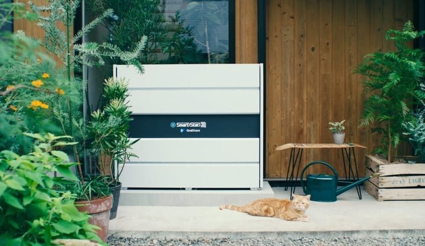 A large gray metal box installed on the back porch of a wood-paneled house. An orange cat lounges on the patio.