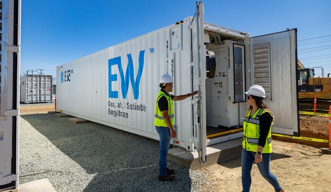 Two people in safety vests and hardhats inspect a large white rectangular metal unit with the letters EW displayed in blue