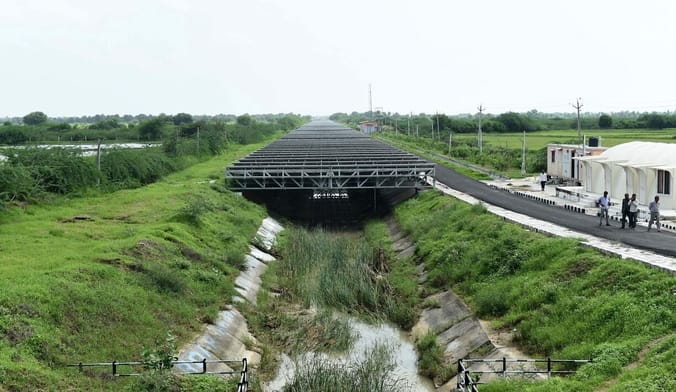 black metal trusses holding solar panels are situated over a grassy canal. Several people walk on a path along the canal.