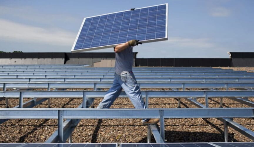 A worker carrying a solar panel in the middle of a large solar array that is under construction