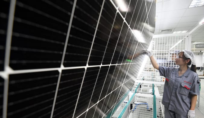 A woman in a factory uniform inspects a large solar panel inside an industrial facility