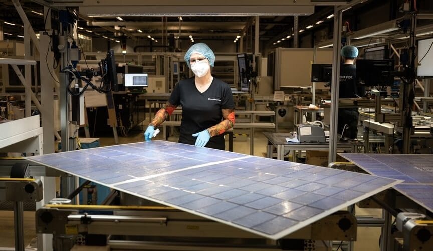 A factory worker with a hair net, face mask, and latex gloves stands next to a large rectangular solar panel