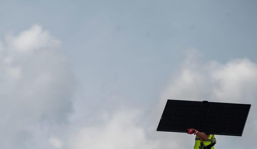 A worker carries a solar panel against a backdrop of cloudy sky