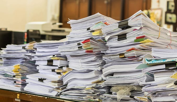several tall piles of paperwork with binder clips and post-it notes stacked on a desk in an office setting