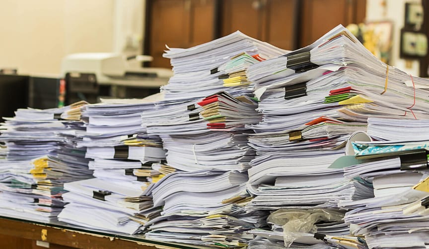 several tall piles of paperwork with binder clips and post-it notes stacked on a desk in an office setting