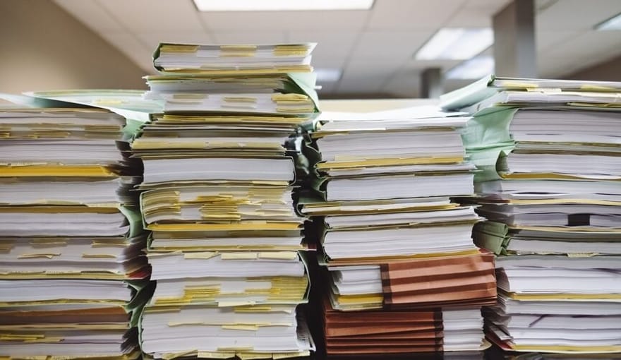 Four tall stacks of paperwork and files in an office setting under fluorescent lights and drop ceiling tiles