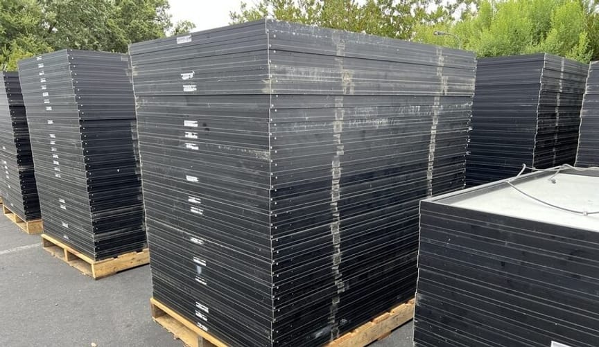 Stacks of black rectangular solar panels on pallets in a parking lot