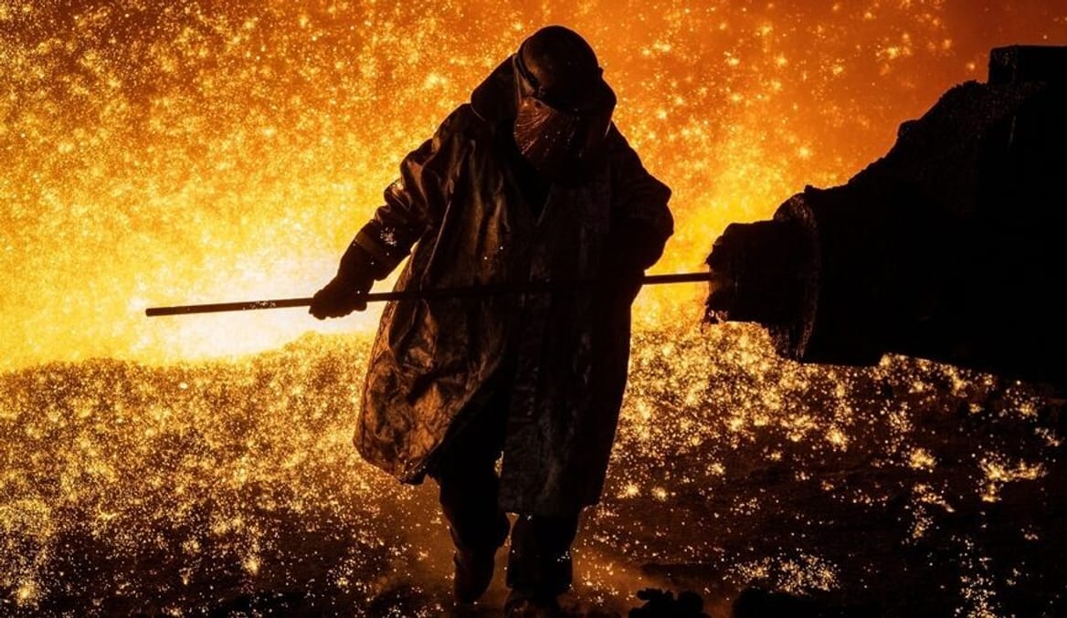 A worker is shown performing labor in a steelmaking facility. Bright orange embers fill the air