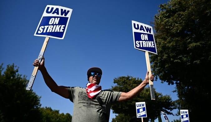 A man wearing a baseball cap, sunglasses and a t-shirt holds up a sign in each hand reading UAW on strike