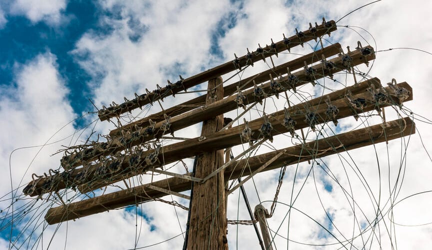 A wooden distribution pole with many tangled power lines against the backdrop of a deep blue sky with wispy white clouds