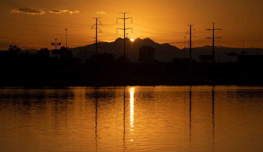 The sun rises behind a mountain range. Powerlines are shown in silhouette. A body of water is in the foreground.