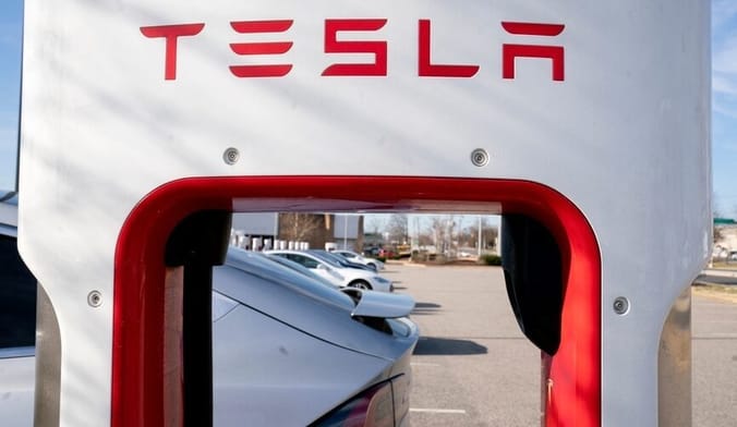 In the foreground is a white EV charger with TESLA in red letters. In the background is a parking lot full of cars