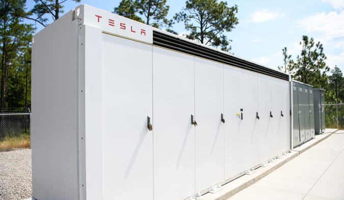Large rectangular row of white metal battery containers bearing the Tesla logo in red letters. The site is bordered by trees.