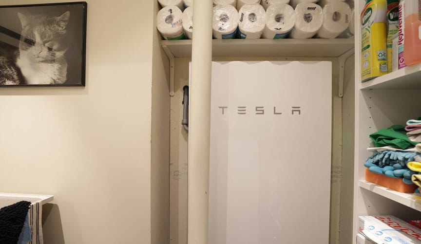 A white Tesla powerwall hangs in a utility room under stacked rolls of paper towels on a shelf and next to a framed cat photo