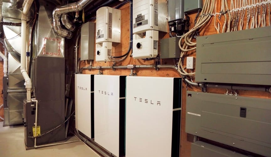 A row of three white Tesla Powerpack batteries on the wall of a garage surrounded by other home equipment