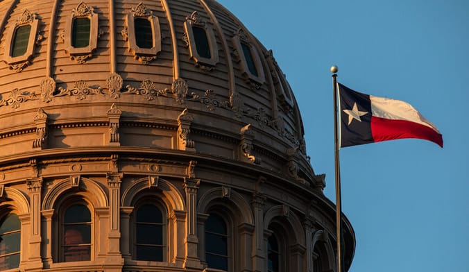 A Texas state flag flies next to the beige stone cupola of the state capitol with a bright blue sky in the background
