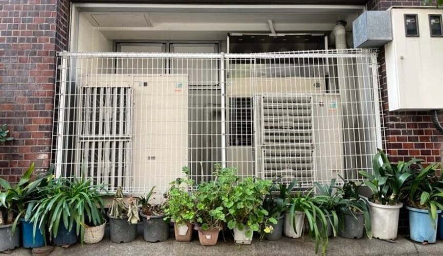Two large heat pumps enclosed behind a metal gate and surrounded by a row of small potted plants
