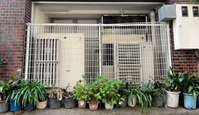 Two large heat pumps enclosed behind a metal gate and surrounded by a row of small potted plants