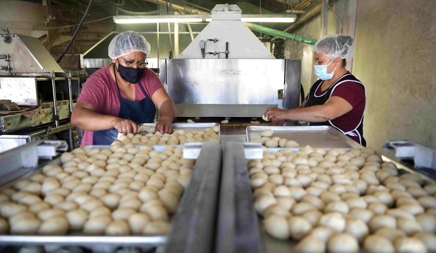 Two women wearing masks and hairnets work next to large trays with hundreds of small dough balls