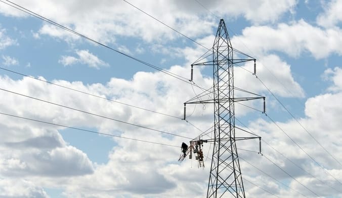 A black transmission tower and power lines against a light blue sky with cotton-y white clouds