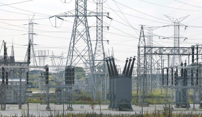 large gray metal transformers and power transmission lines in a fenced outdoor lot