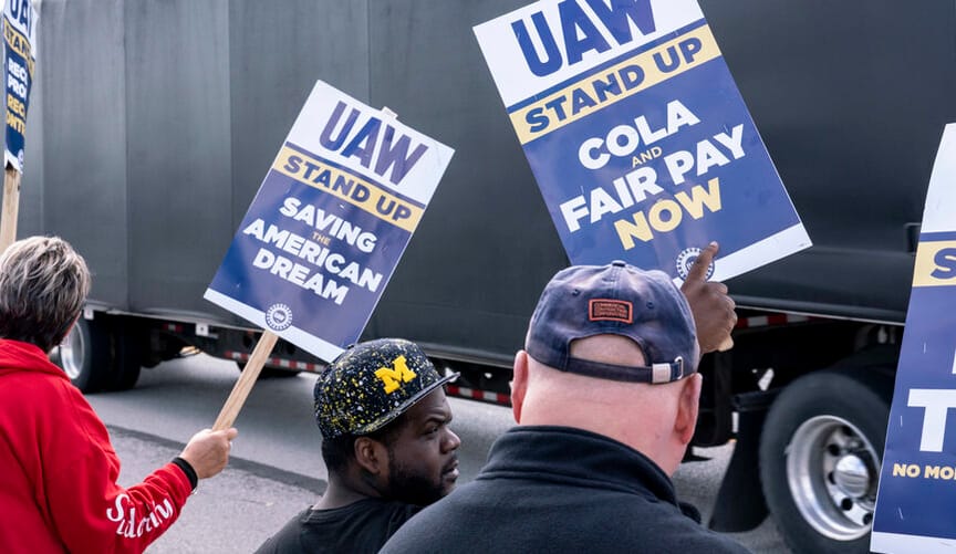 Three people stand on a picket line, holding signs with slogans such as UAW stand up saving the American dream