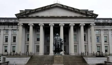The front facade of the US Treasury building in Washington DC. In the foreground is a statue of a man.