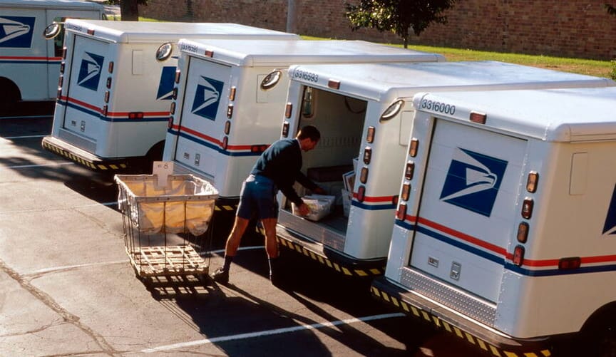 A postal worker loading mail into the back of a mail truck in a post office parking lot