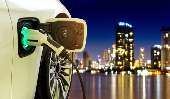 A white EV plugged in to a charger. A city at night is shown in the distant background.