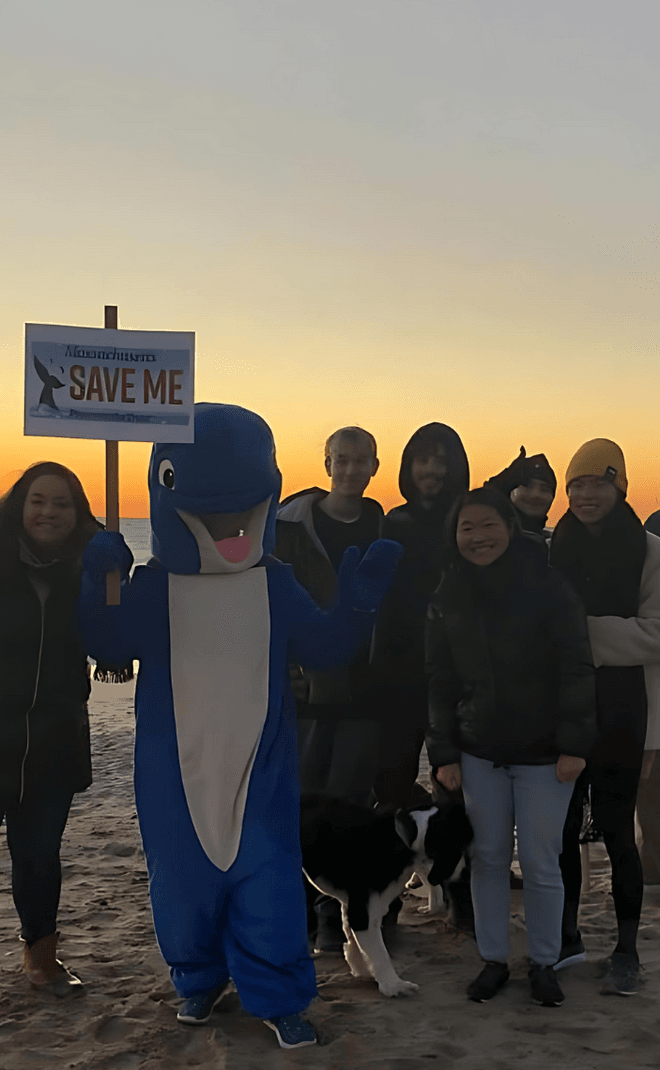 A group of protestors stand on a beach at sunset. One is dressed in a whale costume holding a sign that says SAVE ME.