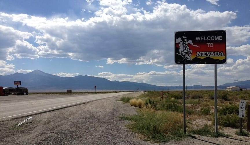 A desert vista with a highway and mountains in background. In the foreground is a road sign that reads welcome to Nevada