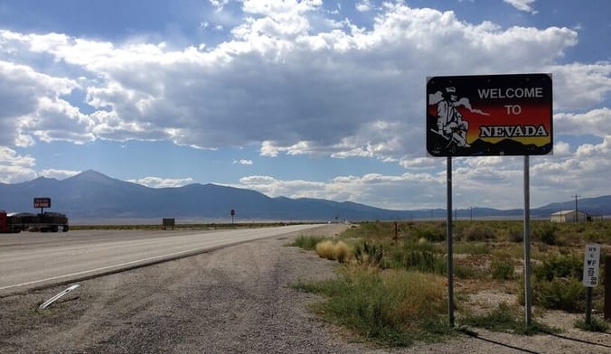 A desert vista with a highway and mountains in background. In the foreground is a road sign that reads welcome to Nevada