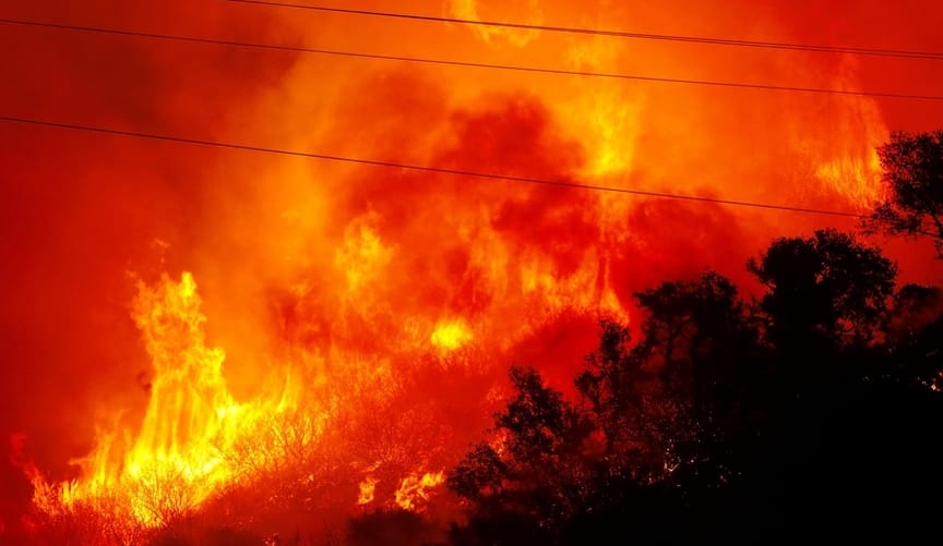 A raging orange and red wildfire engulfs a forested hill. Power lines slash diagonally across the image