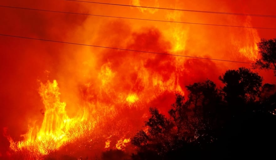 A raging orange and red wildfire engulfs a forested hill. Power lines slash diagonally across the image