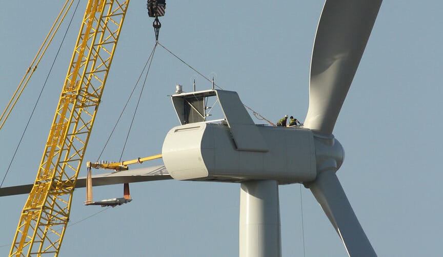 A construction crane and other equipment are attached to the nacelle of a large wind turbine