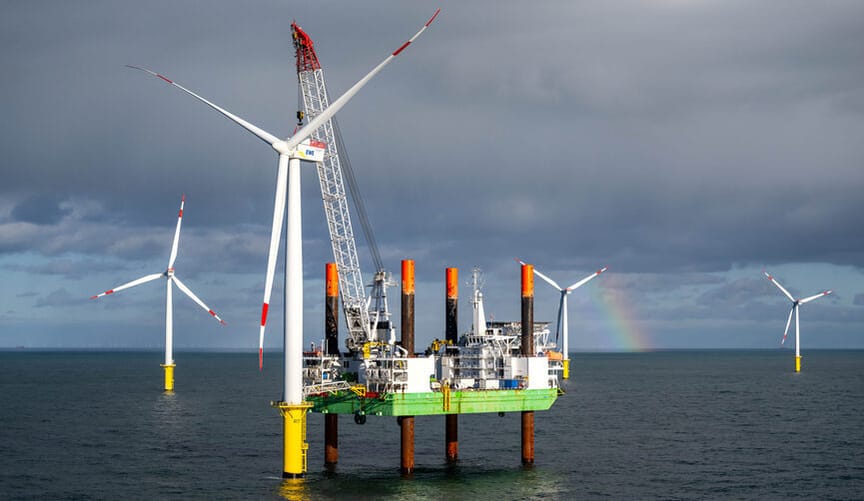 Amid several offshore wind turbines is a green boat laden with construction equipment. Clouds and a rainbow are in the sky.