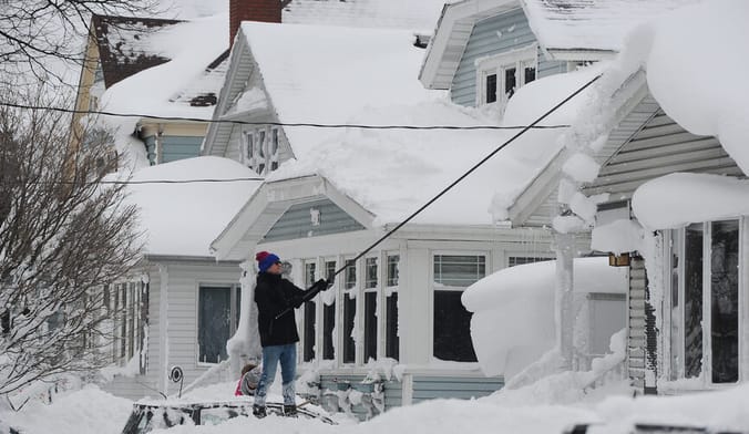 A man in heavy winter clothing scrapes snow off his home's roof with a long device