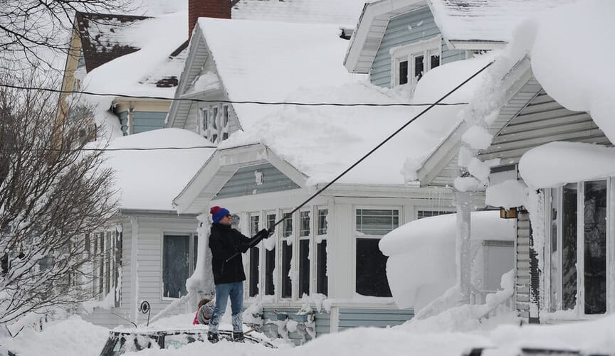 A man in heavy winter clothing scrapes snow off his home's roof with a long device