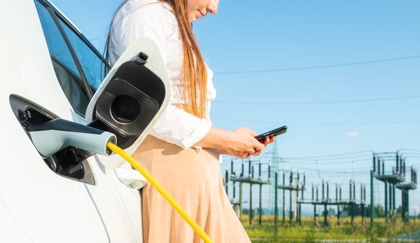 A woman leaning against a plugged-in EV looking a her phone. An electrical grid is seen in the background