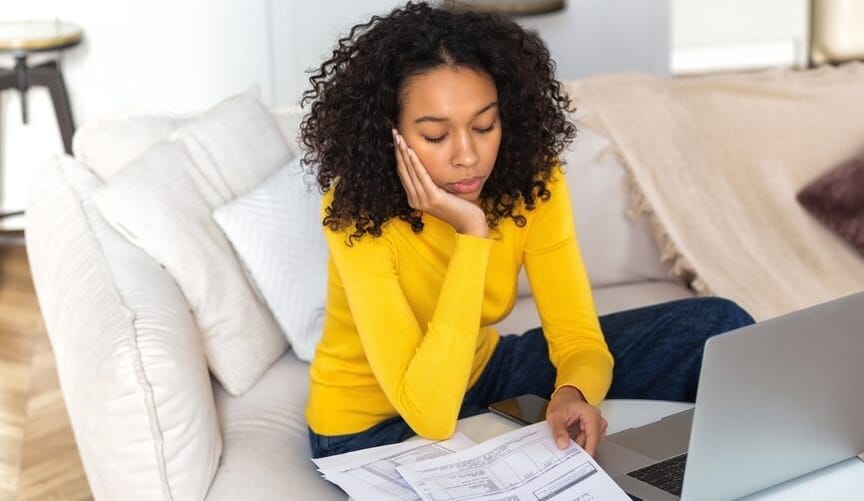 A woman with curly dark hair wearing a yellow sweater sits on a white couch in front of an open laptop looking at bills